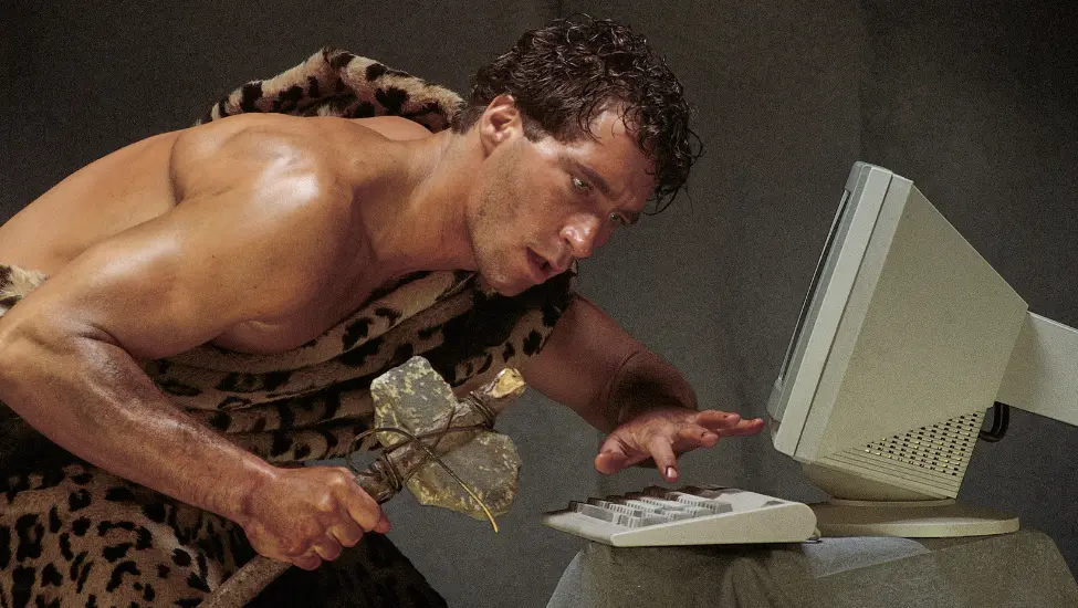 Caveman dressed in animal skin and holding a stone axe trying to use a modern computer