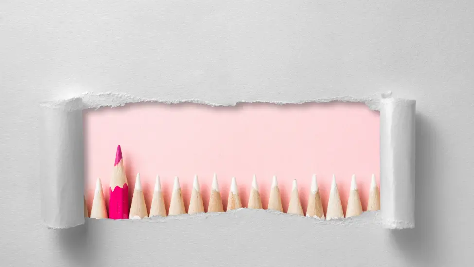 View through torn paper of a row of white pencils with a red leading pencil sticking out