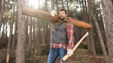 Lumberjack in the forest wearing a plaid shirt holding an axe in one hand and a log on his shoulder with the other hand