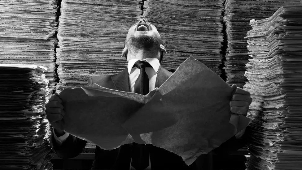 man surrounded by stacks of paper screaming as he tears a piece of paper up