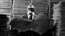 man surrounded by stacks of paper screaming as he tears a piece of paper up