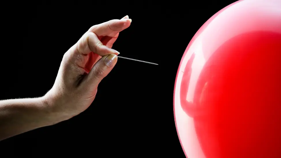 Hand holding a pin to pop a red balloon