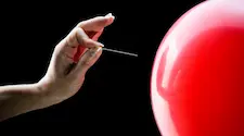Hand holding a pin to pop a red balloon