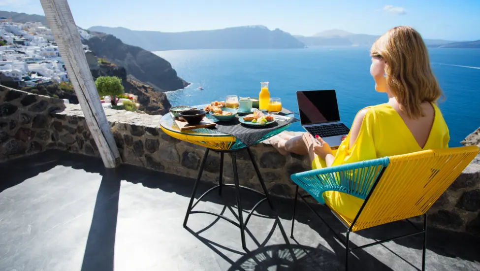 sitting on a patio in the sunshine on vacation looking across at the blue sea while working on a laptop