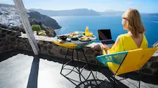 sitting on a patio in the sunshine on vacation looking across at the blue sea while working on a laptop