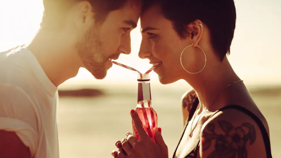 a man and a woman standing head to head and drinking from a shared bottle using strawers